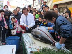 大楽市の会場で、市長が大きなマグロを解体しており、その様子を沢山の市民の方が集まって見ている写真