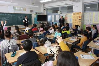 教室で手を挙げ授業を受けている子供達を、時計の下に立ち見学している市長を教室後ろから全体を写した写真