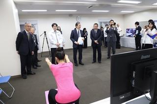テレビが設置されている前でピンク色のティーシャツを着た女性がスクワット運動をしており、市長とKDDI総合研究所の方々がその様子を見ている写真
