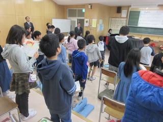 音楽室にて先生のピアノの伴奏に合わせて子ども達が歌を歌っており、市長がその様子を前方の入り口付近で見ている写真