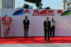 大井朝市の横断幕のかかっている舞台の上で市長がスタンドマイクの前に立って話をしており、ぶじみんと関係者の男性が舞台の後ろに立っている写真
