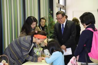 オレンジのバンダナをしている女性と女の子が机を挟んで向かい立っており、バンダナの女性が女の子に何か書いており、その様子を市長が笑顔で見ている写真