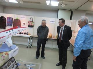 作品展の展示会場に、手作りの洋服やバックが展示されており、手前には飛行機のような物が展示されていて、市長と、年配の男性2名が、飛行機のような作品に注目してみている写真