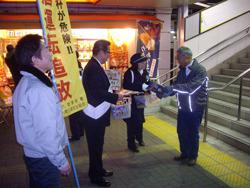 交通事故防止運動に参加されているグレーのジャンパーを着た男性が「飲酒運転追放」の文字が書かれているのぼり旗を持っており、その奥で、駅の階段から降りて来た市民の男性に、市長と関係団体の女性がチラシを手渡している写真