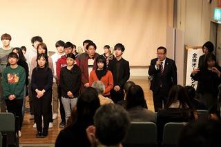 舞台前に並んでいる人の横で市長がマイクで挨拶をしている写真