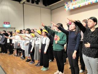 舞台上でたくさんの人達が並び右手を上にあげている写真
