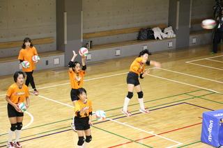 5名のオレンジと黒のユニフォーム姿の女子バレー選手たちがサーブを打っている写真