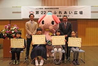 「第22回 ふれあい広場」と書かれている横断幕の前で、表彰を受けた児童、生徒4名が表彰状を手に持って、椅子に座っており、市長とふじみんと関係者の男性と写っている記念写真