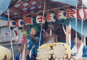 苗間はやしで木組みの屋台の上で太鼓や笛を吹く男性達とひょっとこのお面をつけた男性が映っている写真