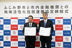 市長と眼鏡をかけてブルーのネクタイを身に着けている市内金融機関代表の方が締結書を持って横並びで立っている写真