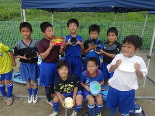 サッカーのユニフォームを着た少年たちが9人ほど並んでいて、それぞれ空になったお弁当箱を見せている写真