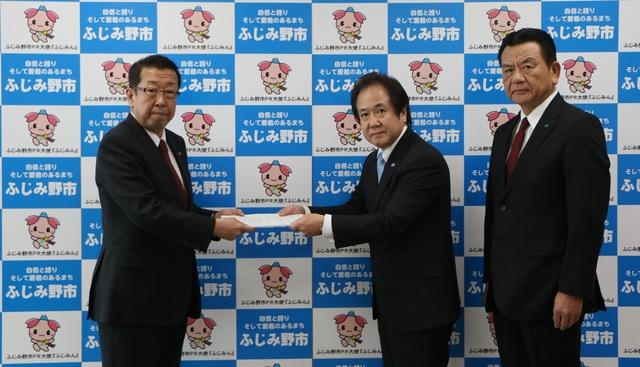 スーツを着た男性が市長に一次答申書を手渡し、また、その後ろに一人男性が立っている写真