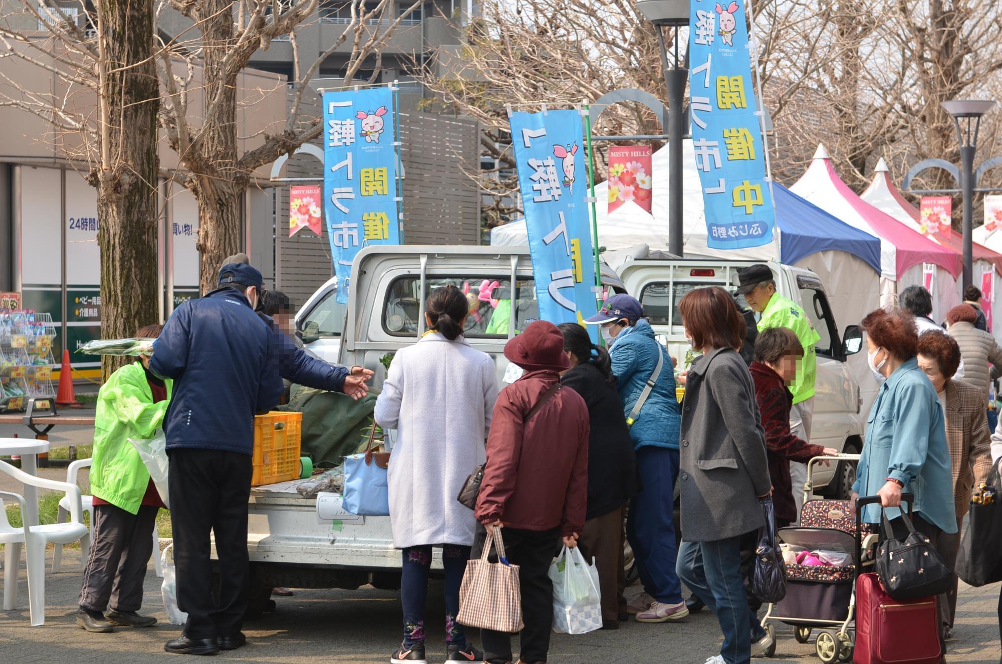 軽トラ市開催中と書かれたのぼりが軽トラの横にあり人々が軽トラの周りに集まっている様子の写真
