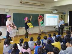 小学生の児童の前で、「認知症」、「老化」、等のプレートを首から下げた講師の方々が認知症について分かりやすく目の前で表現して説明をしている様子の写真