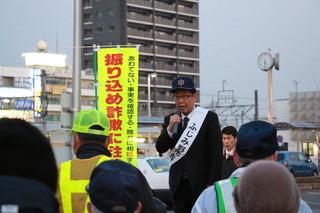 帽子を被り襷をかけた市長がマイクを持って市民の前で街頭演説をしている写真