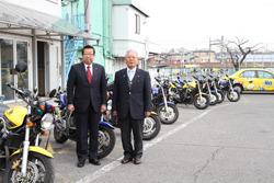 沢山の教習用車両のバイクが並んでいる前で、市長とセイコーモータースクールの関係者の男性と写っている写真