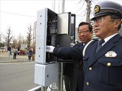 交差点の電信柱に着いている信号機の制御機の点灯を、市長と警察官の男性がボタンに手を伸ばして点灯しようとしている様子の写真