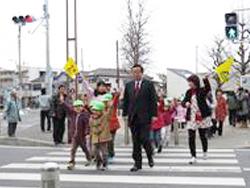 横断歩道用の信号機が青に点灯し、横断歩道を園児と先生が手を挙げて歩いて渡っており、市長も一緒に歩いている写真