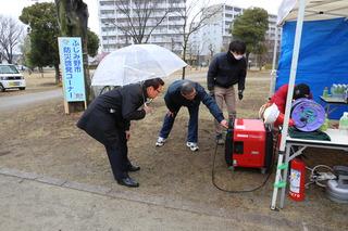 防災啓発コーナーで発電機が動いてる様子を男性スタッフから傘をさして説明を受ける市長の写真