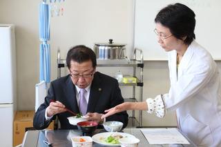 調理された料理を試食する市長に女性スタッフが説明している写真