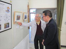 歴史民俗資料館友の会作品展示会の会場の壁に展示されている額に入っている写真のような作品を、白髪の男性が指を指しながら、市長に説明をしている様子の写真