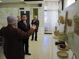 手作りのかごの作品がボードや机に展示されており、帽子を被った男性が説明をしているのを市長が聞いている様子の写真