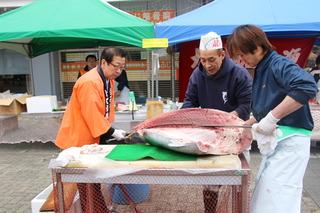 さくらまつりのココネ広場にてマグロの解体ショーにオレンジ色の法被姿の市長も参加している写真