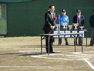 野球大会の開会式で市長が挨拶している横側の机に優勝カップが並べられており、審判員たちが立っている写真