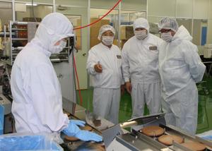白い防護服を着て見学をしている市長と関係者が、工場内で茶色で丸いお菓子が焼きあがり、レーンに並んでいる様子を、市長が工場の職員の方から説明を受けている写真