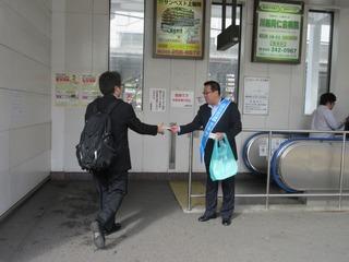 駅のホームに入って行く、リュックを背負った男性にチラシを手渡している市長の写真