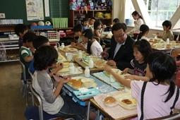 給食の時間、机を向かい合わせにして市長が子ども達の間に座って、横に座っている女子児童に話しかけながら、楽しそうに給食を食べている写真
