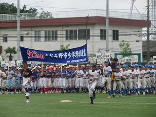 少年野球大会の各チームがユニフォームを着て、チームのプラカードを持って整列しており、代表の選手2名が旗を持って行進している様子の写真