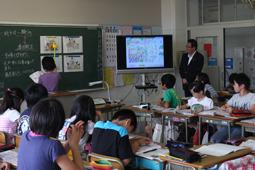 教室前のテレビに映し出された画面を見ながら黒板のプリントを指している生徒の後ろ姿を見ている市長と子供達の全体を写した写真