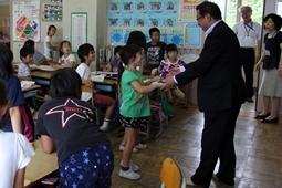 教室前に立つ市長と緑のシャツを着た女の子が握手をしている所を周りの児童、先生が見ている写真