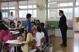 青いシャツを着た男の子が教室の後ろにいる市長に質問をしている様子を周りの児童達が振り返って見ている写真