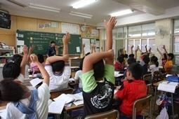 教室の左斜め後方から児童2名が右手を上にあげ左手で右手を支えている後ろ姿をアップにして全体を写した写真