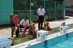 黄色の水泳帽をかぶりピンクのビート板をもったり、その上で横になったりとプールサイドで休憩している子供達に話しかけている市長と関係者の人の写真