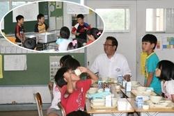 西小学校にて給食時間に生徒が配膳している様子を給食の配られた机から見学する市長の写真