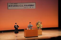お花の飾られた演題で野口 健さんが講演をしている横で女性が手話通訳をしている写真