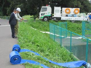 道路から河川に水色のホースが2本伸びており、関係者の男性が市長に説明をしている写真