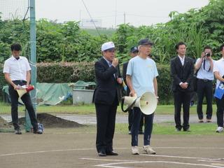 早起き野球大会のグラウンドで、関係者の男性が拡声器を持ち、その横で市長が拡声器のマイクを使い話をしている写真