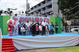 おおい祭りの会場の舞台で、市長が話をしており、市長の横には手話の同時通訳者の女性が手話をしており、その後ろにはふじみんと関係者の方々がパイプ椅子に座ったり、立って話を聞いている写真