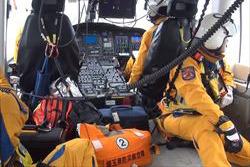 ヘリコプターの中で、ヘルメットにオレンジ色の制服を着用した救命士が運転席に1名と背中合わせに座っている1名の写真