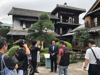 福岡河岸記念館の庭で、市長がテレビ局の方の話を聞いており、撮影をする関係者の方々がその様子を見守っている写真