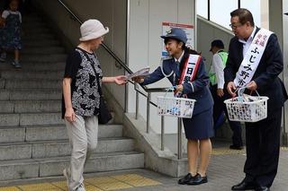 婦人警官の服を着ている重田 彩さんと市長が駅の階段から降りて来た帽子を被った女性にチラシを手渡ししている写真