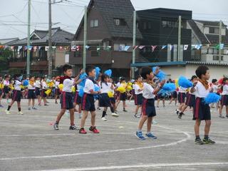 低学年の児童達が運動場に広がって、青いボンボンを両手で持って振りながら、ダンスを踊っている写真