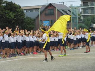 黄色いチームの団長が黄色い団旗を振って、黄色いチームの児童は団長に合わせて両手を頭の上に挙げて手を叩いて応援して盛り上がっている写真