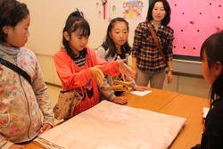 小学生の女子児童が机に集まっており、ピンクの洋服を着た女の子が両手の間で木が浮いている手品を見せている写真
