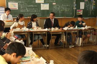黒板の前に並べられた机で子どもた達と一緒に楽しそうに給食を食べている市長の写真