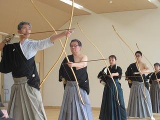 袴を着た方々が弓道場に並んで、弓を構えて的に向かって打とうとしている写真